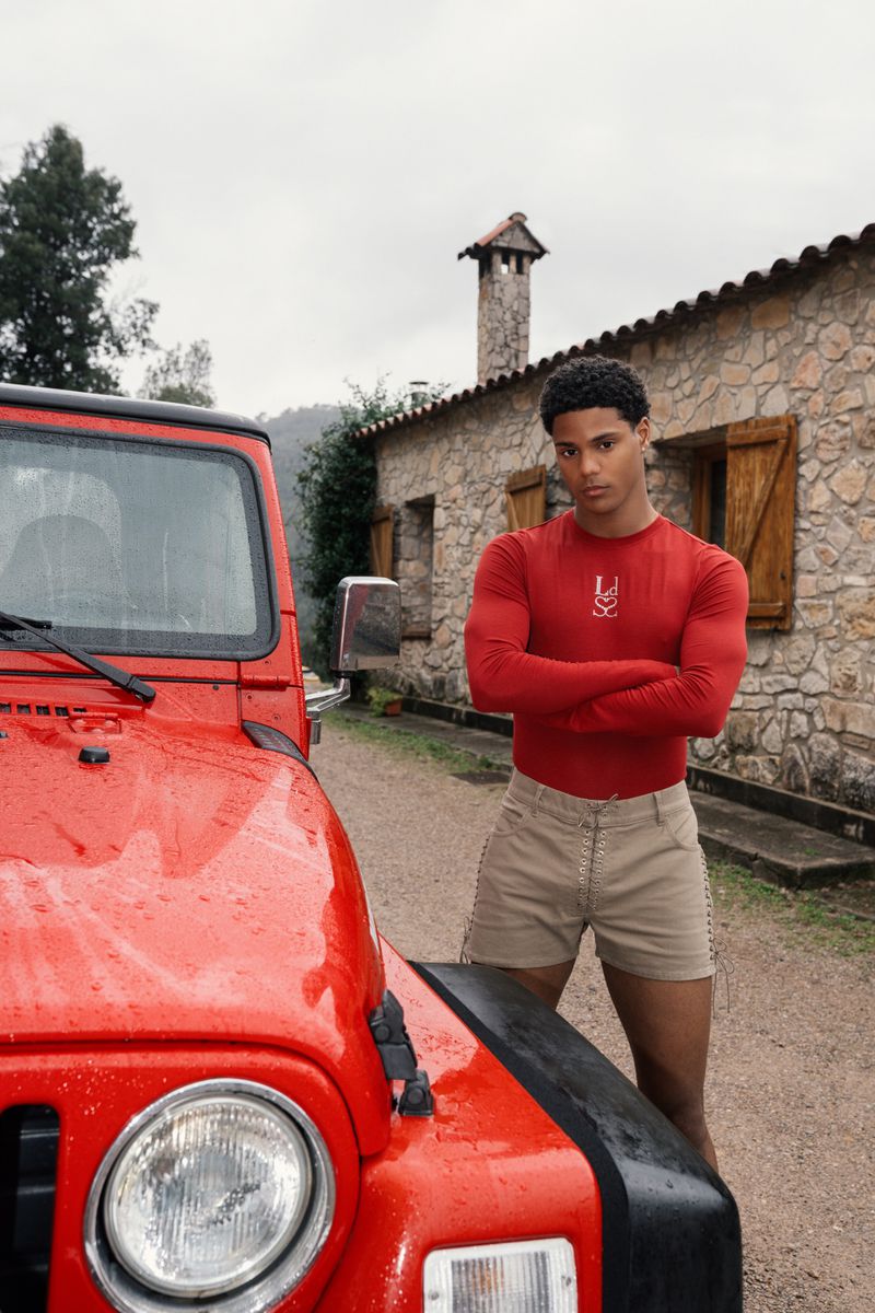 Portrait of a male model in red long sleeve shirt and khaki shorts, posing with a red Jeep against a stone house backdrop.