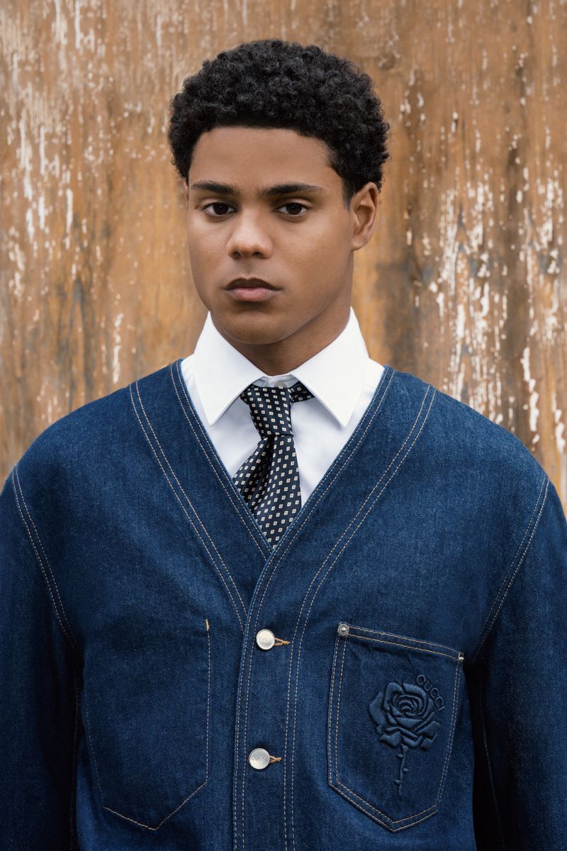 Portrait of a young man wearing a blue denim cardigan over a white dress shirt and patterned tie, posing against a rustic wooden wall.