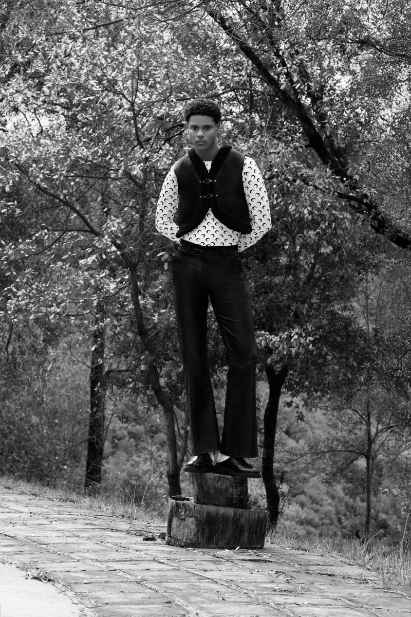 A male fashion model poses outdoors on a tree stump wearing dark trousers, a patterned shirt, and a black vest against a blurred forest background.