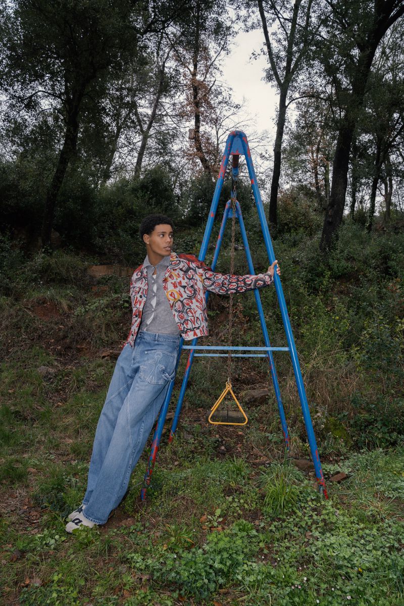 Young adult male poses with vintage style in an outdoor setting with a metal swing set, wearing denim and a patterned jacket.