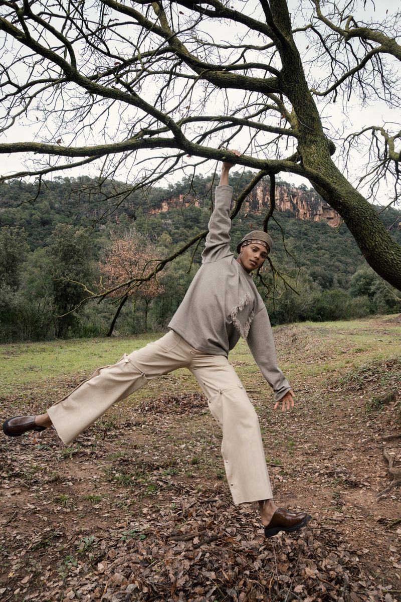 A man in tan clothing hangs from a tree branch in a field with trees and hills in the background on a cloudy day.