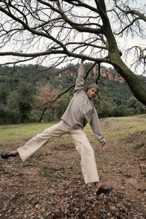 A man in tan clothing hangs from a tree branch in a field with trees and hills in the background on a cloudy day.