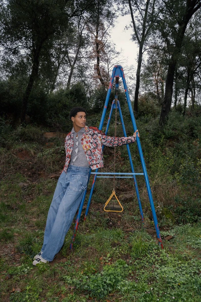 Young man leaning against a blue metal swing set frame outdoors in a wooded area wearing a patterned jacket and loose blue jeans.