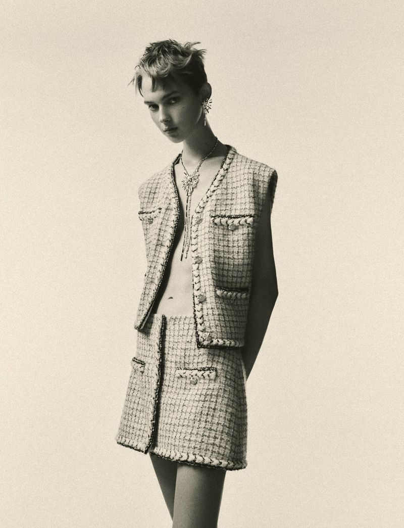 A slender fashion model poses in a sleeveless tweed vest and skirt against a neutral backdrop, showcasing a stylish, vintage-inspired look.