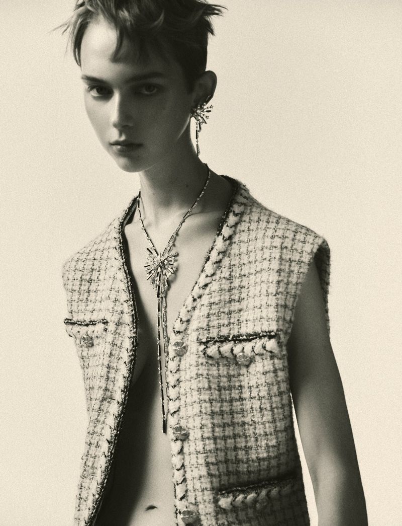 Monochrome studio portrait of a stylish adult woman with short hair wearing a tweed vest and ornamental jewelry, set against a neutral backdrop.