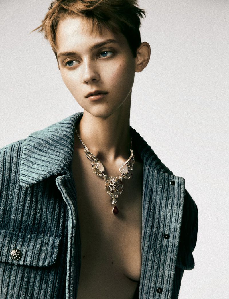 A young adult woman with short brown hair poses indoors wearing a turquoise corduroy jacket and statement necklace against a neutral background.