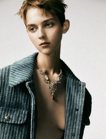 A young adult woman with short brown hair poses indoors wearing a turquoise corduroy jacket and statement necklace against a neutral background.
