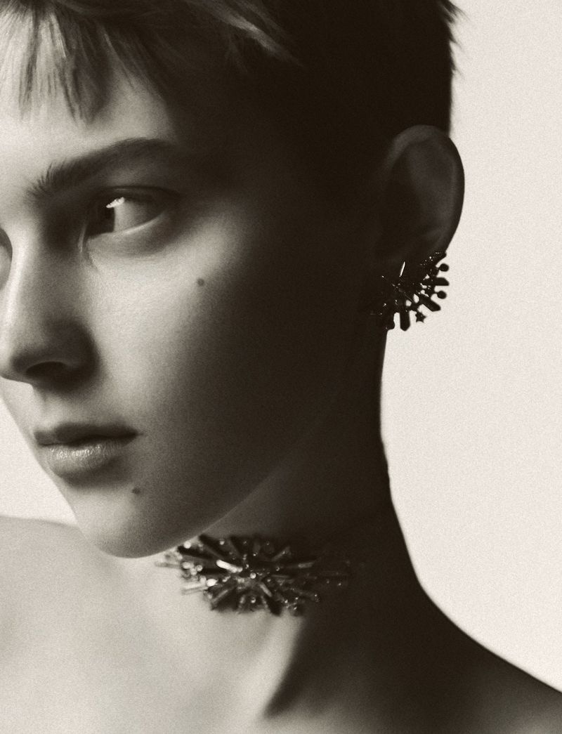 Close-up portrait of a woman with short hair wearing a statement choker necklace and stud earrings against a neutral backdrop.