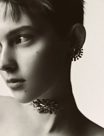 Close-up portrait of a woman with short hair wearing a statement choker necklace and stud earrings against a neutral backdrop.