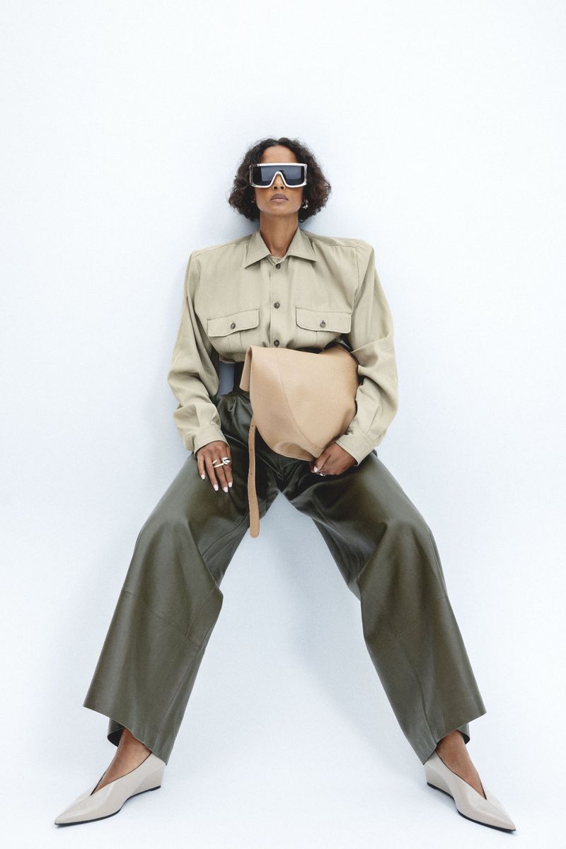Fashion shot of a Black female model in beige shirt and wide-legged green pants, holding a beige bag, posing in front of a white wall.