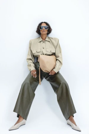 Fashion shot of a Black female model in beige shirt and wide-legged green pants, holding a beige bag, posing in front of a white wall.
