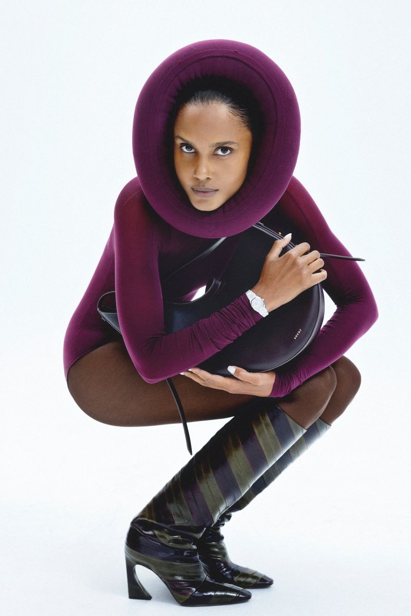 Woman in plum bodysuit and striped boots poses holding a black bag in a studio. White background. Fashion editorial shot.