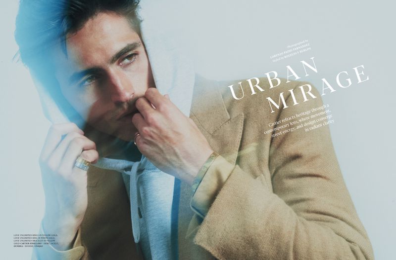 Man poses indoors wearing a hoodie and jacket, highlighted by bright lighting, showcasing a contemporary fashion style and jewelry.