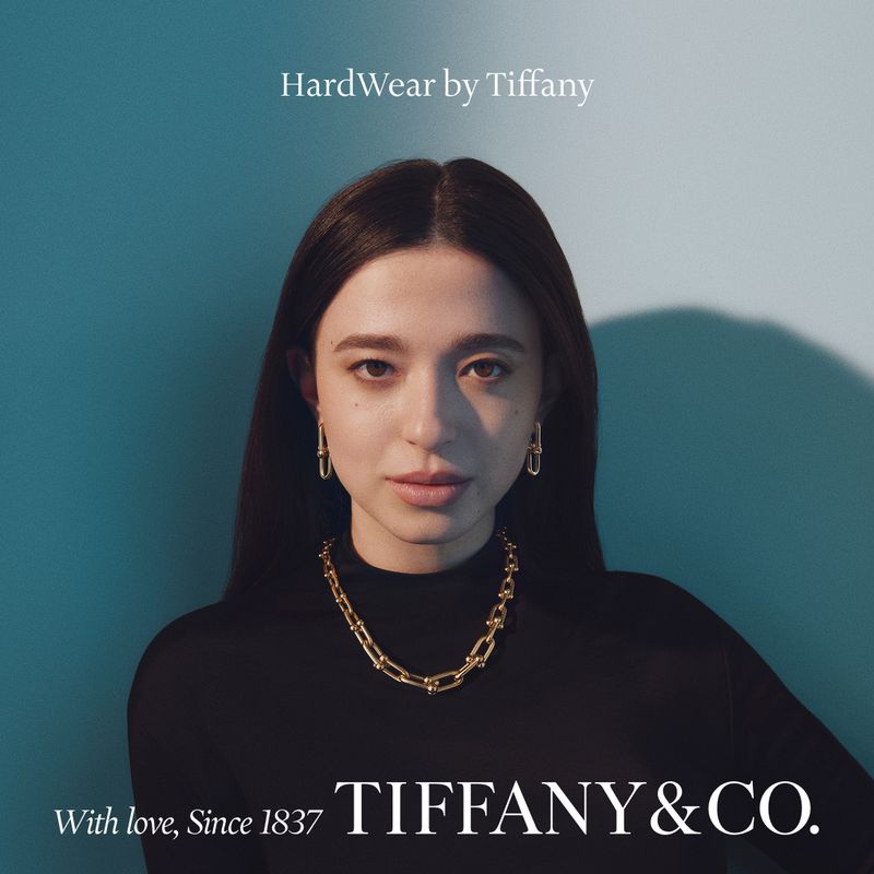 Portrait of a woman wearing gold chain jewelry and black top against a teal background for Tiffany & Co. campaign.