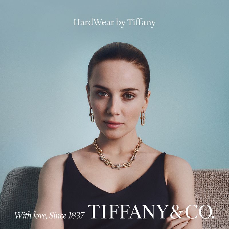 Portrait of a woman wearing gold chain jewelry and a black top against a solid light blue background for Tiffany and Co.