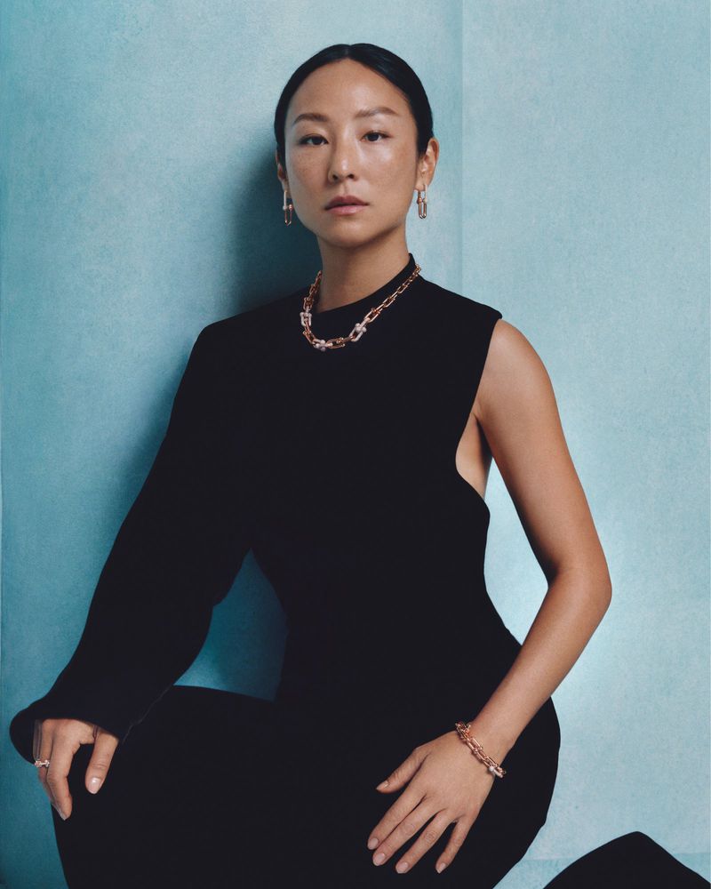 Portrait of Greta Lee wearing gold chain jewellery and a black dress against a textured blue wall for Tiffany & Co.