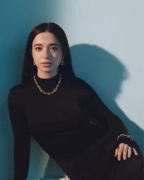 Portrait of a woman wearing gold chain jewelry and a black top against a pale blue background for Tiffany and Co.