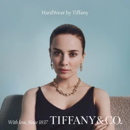 Portrait of a woman wearing gold chain jewelry and a black top against a solid light blue background for Tiffany and Co.