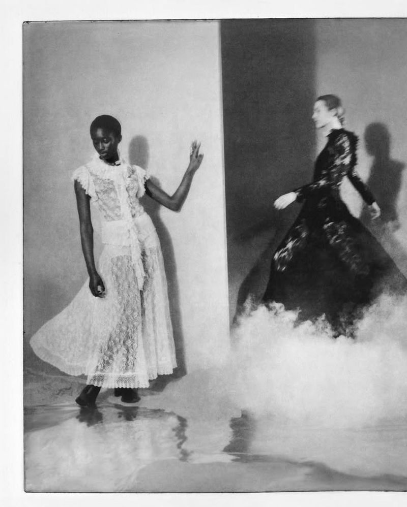 Two women in elegant lace dresses pose in a studio with textured lighting, creating a dramatic black-and-white fashion editorial shot.