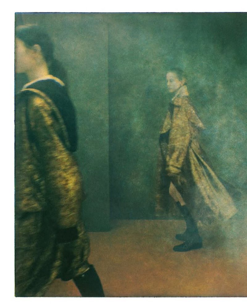Two young women are walking indoors, one in a gold dress with a black hood, and another in a gold trench coat and black boots.