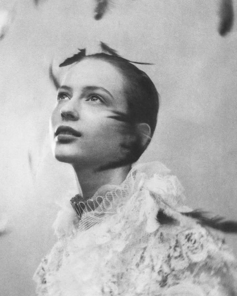 Black-and-white portrait of a woman looking up, adorned with feathers and wearing a lace collar, captured in a studio setting.