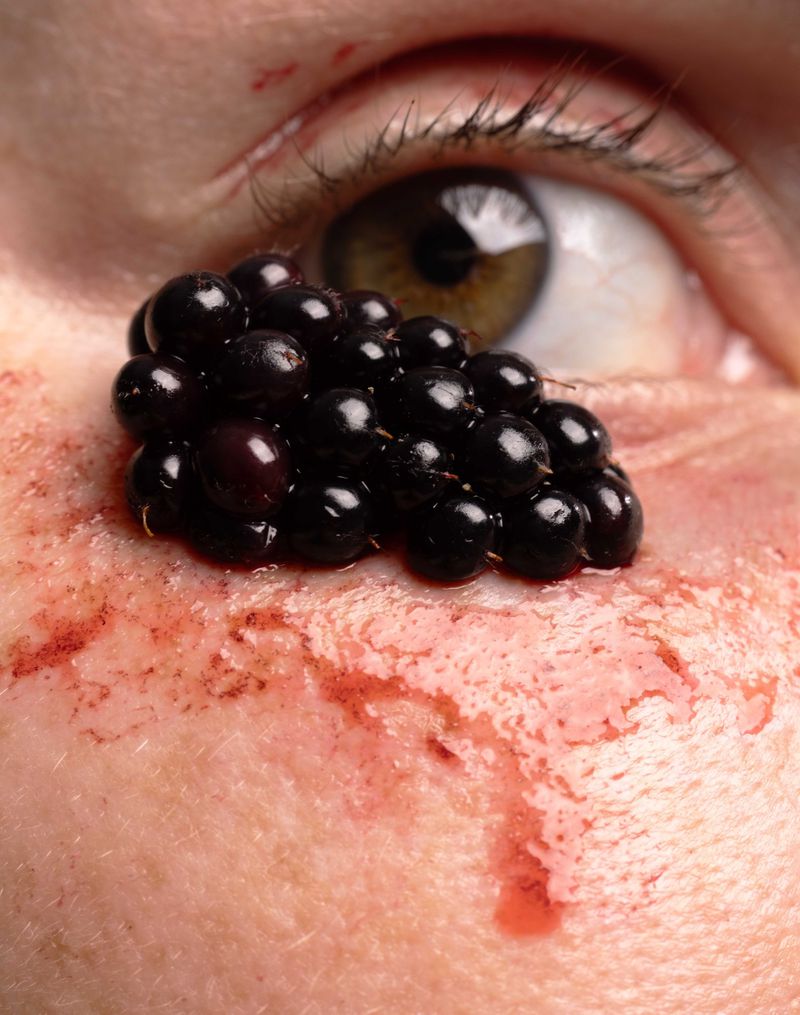 Close-up of an eye and part of a face covered in blackberries and a red, liquid substance, creating a surreal and unsettling image.