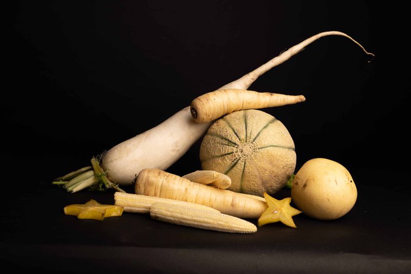 Still life with assorted melon, star fruit, radish, corn, and root vegetables on a dark background showcasing culinary ingredients.