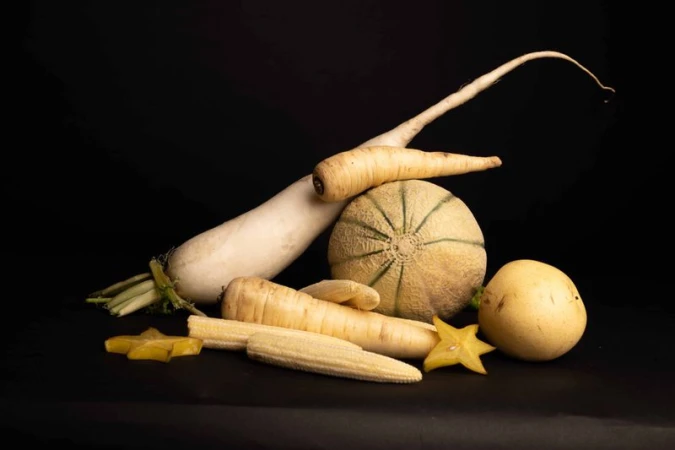 Still life with assorted melon, star fruit, radish, corn, and root vegetables on a dark background showcasing culinary ingredients.