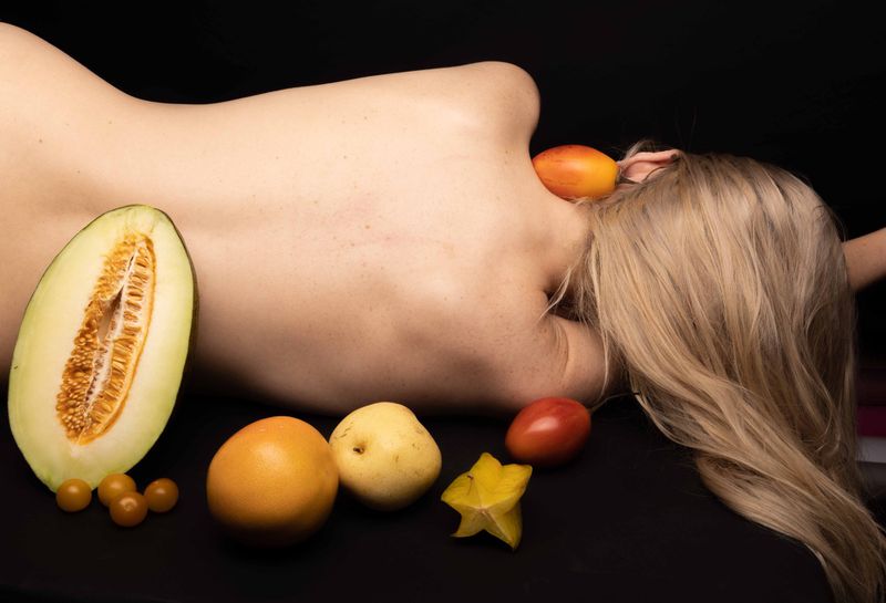 Nude back of a blonde woman surrounded by colorful assortment of melon slices, starfruit, tomatoes, pears, and yellow plums on a black backdrop.