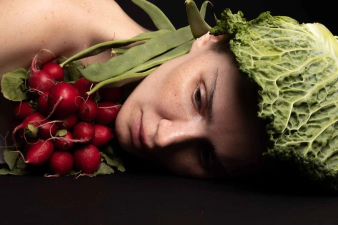 Eye-level shot of a woman's face adorned with fresh vegetables: radishes, green beans, and a cabbage on a black background.