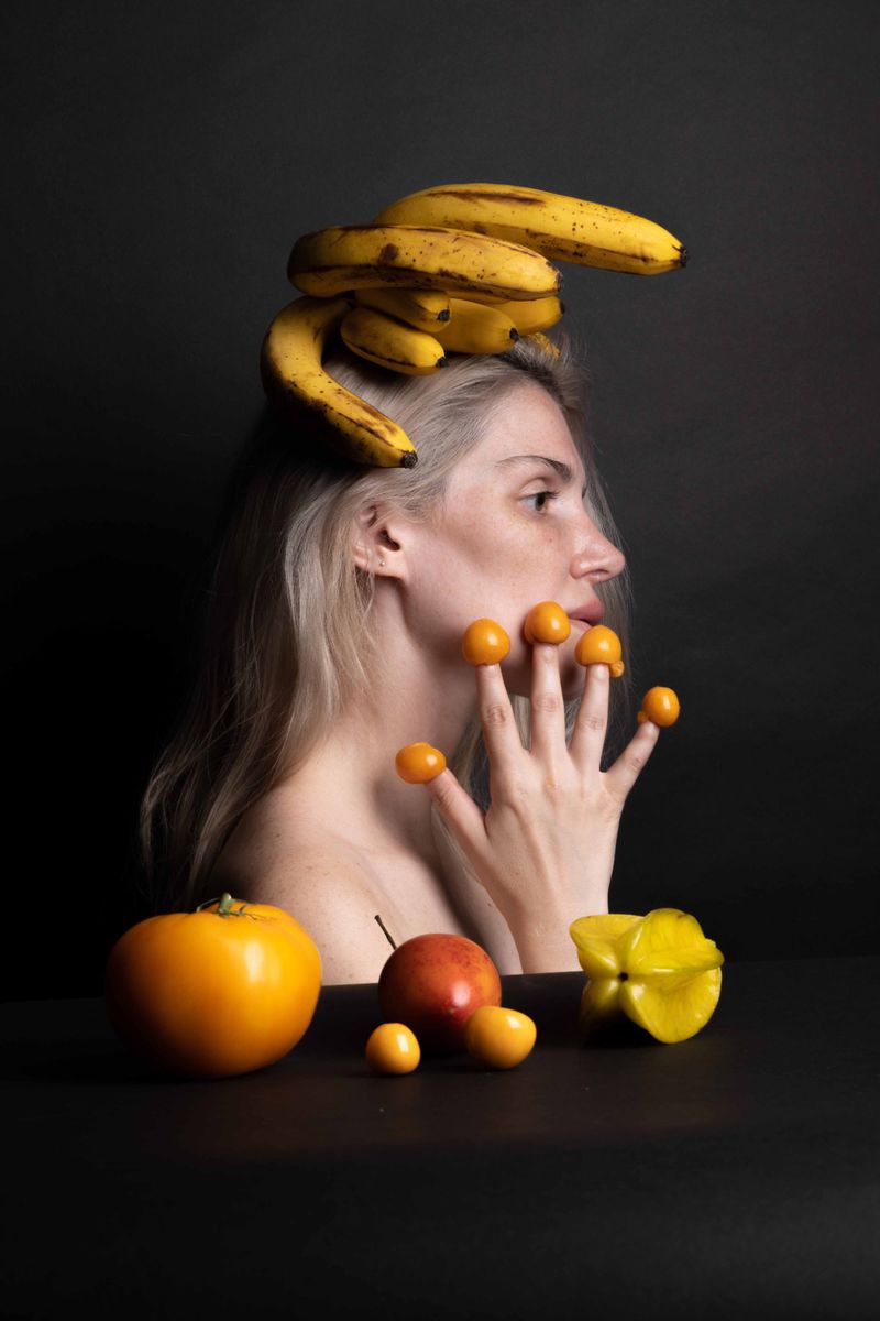 Conceptual portrait of a woman with fruit. She has bananas on her head and small oranges on her fingers. A tomato and carambola sit below.