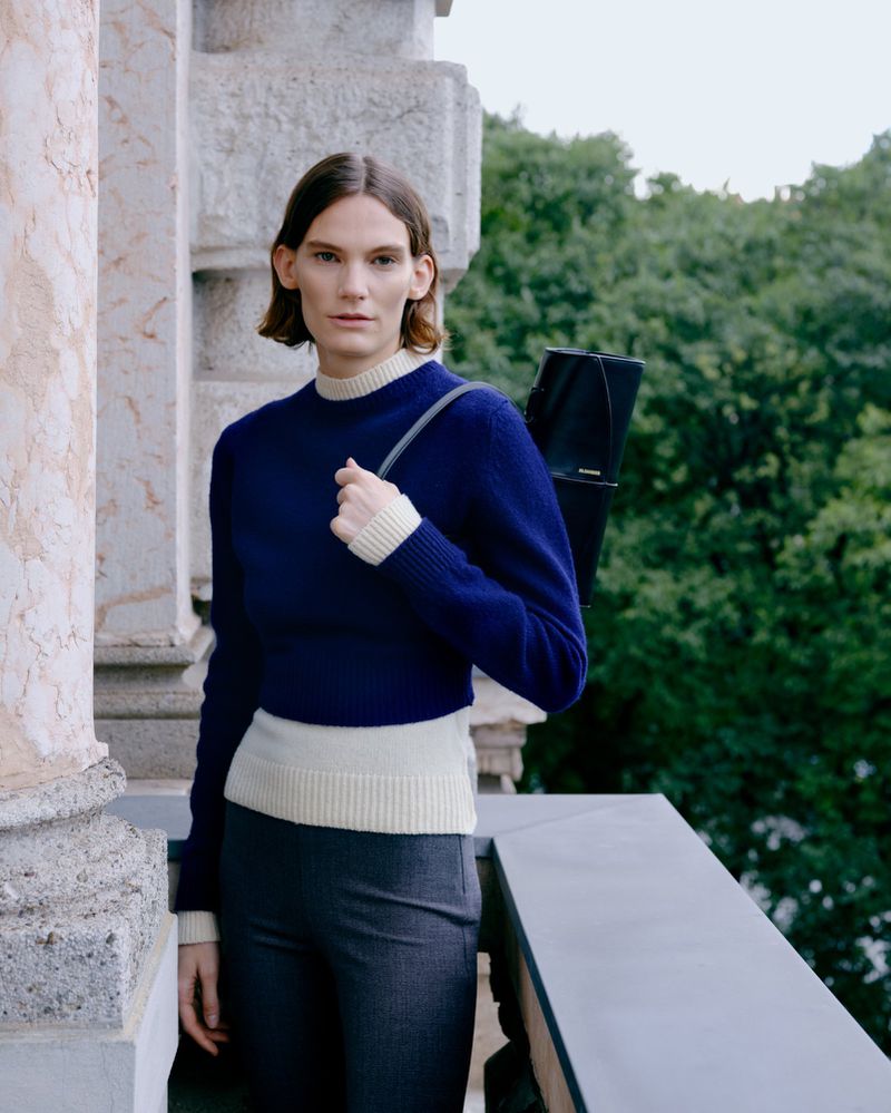 Woman in blue sweater and grey pants carrying black tote bag outdoors, posing against a stone railing with trees in the background.