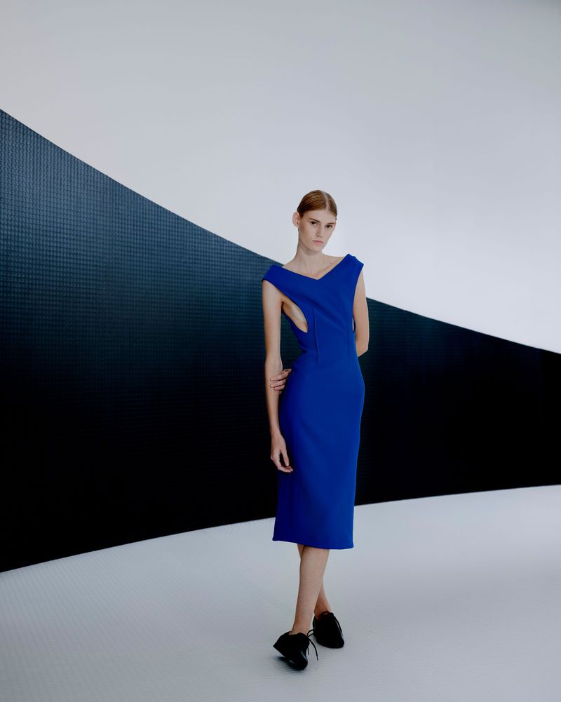 Full-shot of a female model in a blue, midi-length dress with geometric cut-outs, posing against a geometric black and white backdrop.