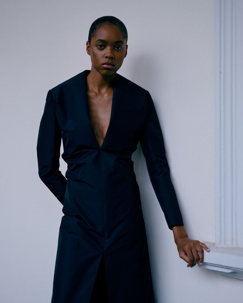 Portrait of a Black woman wearing a dark, deep V-neck dress, indoors against a plain white wall with light from the side.