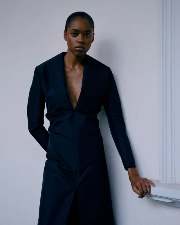 Portrait of a Black woman wearing a dark, deep V-neck dress, indoors against a plain white wall with light from the side.