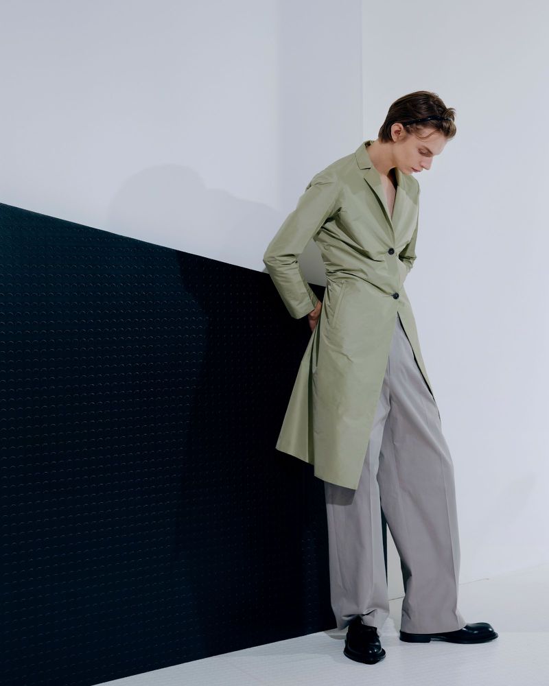 Fashionable adult wearing a long, sage coat and wide grey trousers standing in a photo studio against black and white geometric backdrop.