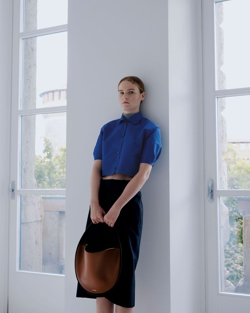 Indoor portrait of a woman in a blue top and black skirt, holding a brown leather purse, near a window with natural light.