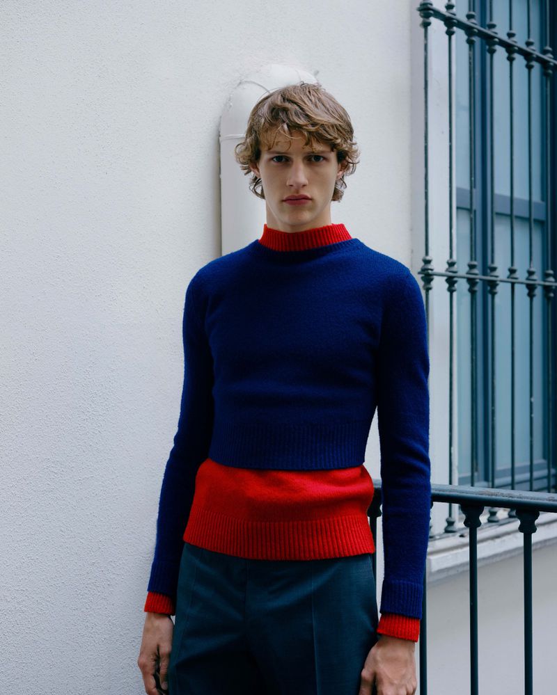 Blonde man in blue sweater and green pants poses in front of white wall and dark window for a fashion shot.
