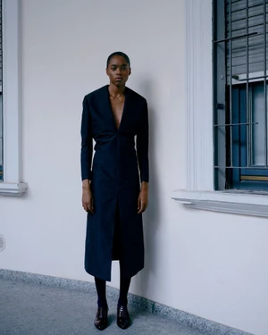 Model wearing a dark structured long dress and dark shoes standing against a plain white wall outdoors.