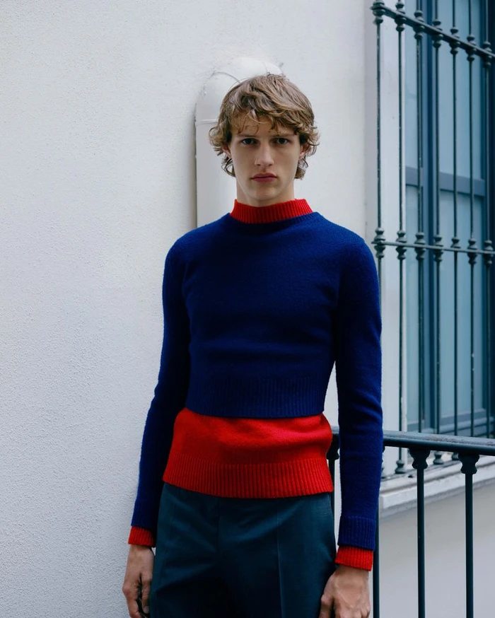 Model wearing a blue knit sweater with a red collar and hem standing against a white building exterior.