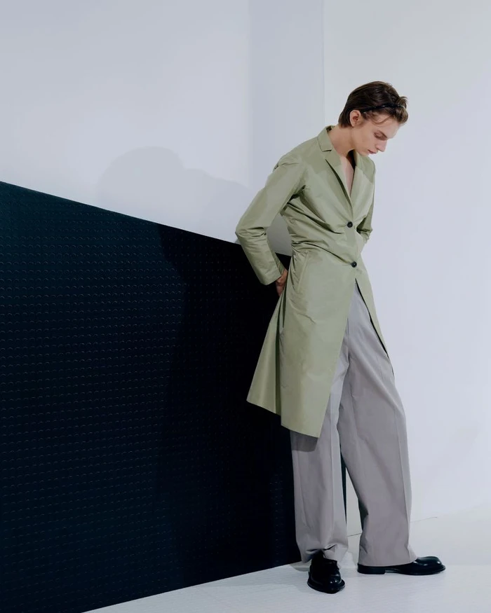 Model posing in a tailored sage green coat and wide-leg grey trousers against a minimalist black and white wall.