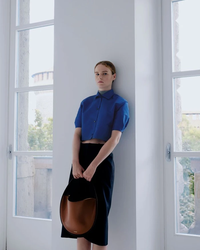 Model in a blue short-sleeve shirt and black skirt holding a brown leather handbag, leaning against a white pillar between tall windows.
