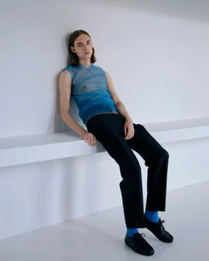 Model with long hair wearing a blue sheer sleeveless top and black trousers leaning against a white studio wall