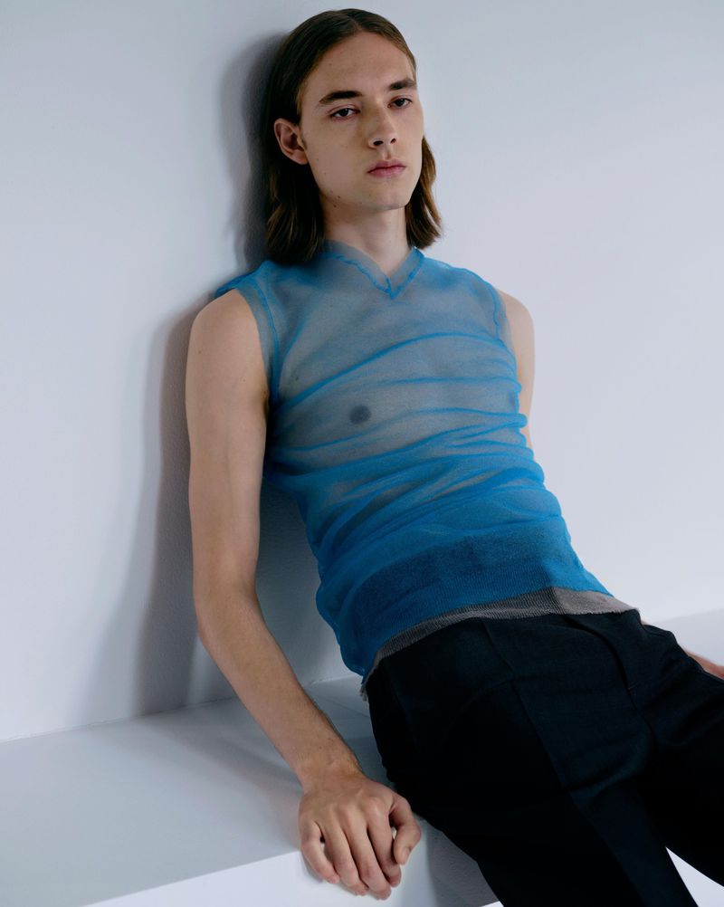 A young male model with long brown hair wearing a sheer blue sleeveless top and dark trousers leaning against a white studio wall.