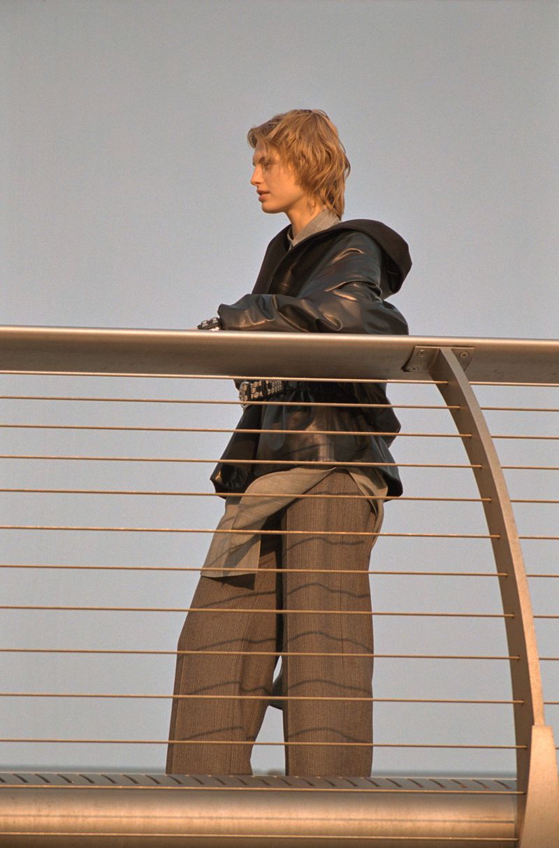Adult with blond hair in oversized grey striped pants and black jacket leaning on silver railing against a light grey sky background.