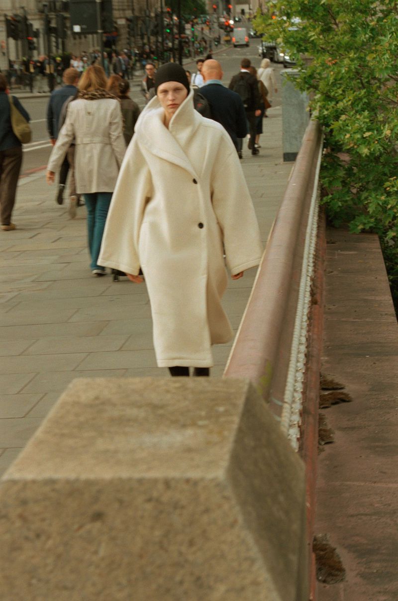 A woman in a long, light-colored coat walking on a sidewalk with other pedestrians, with greenery on the right and urban buildings in the back.