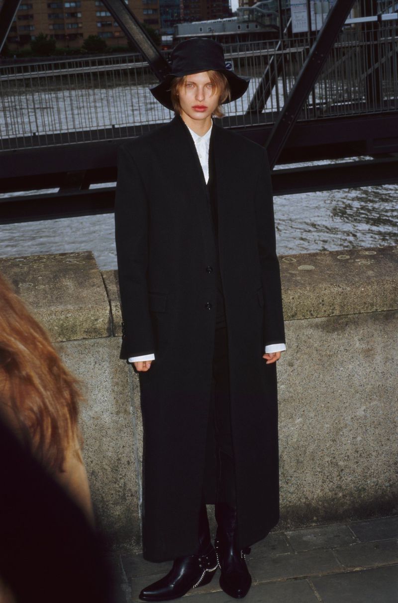 A lone model stands against an urban backdrop, sporting a black bucket hat, a long black coat, and stylish black boots.