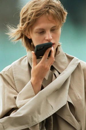 A young adult with short blonde hair holds a smartphone to their face wearing a beige trench coat in an outdoor setting.