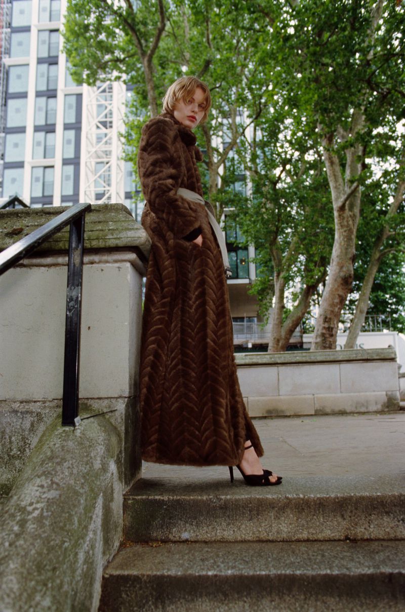 A woman in a brown herringbone coat and high heels stands on concrete steps outside with a blurred background of buildings and trees.