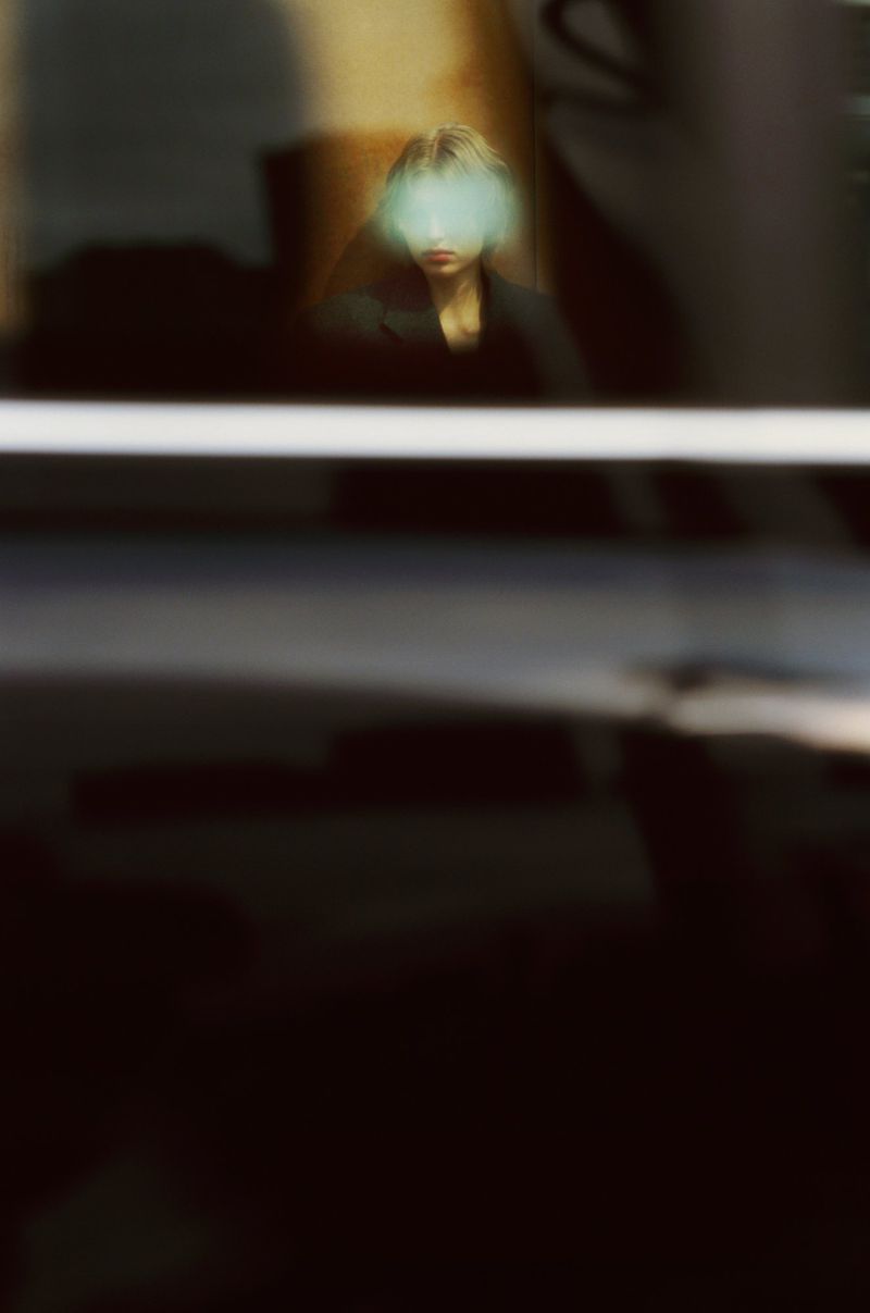 Abstract portrait photograph of a woman with short blond hair in a dark coat, shot through a reflective glass surface indoors.